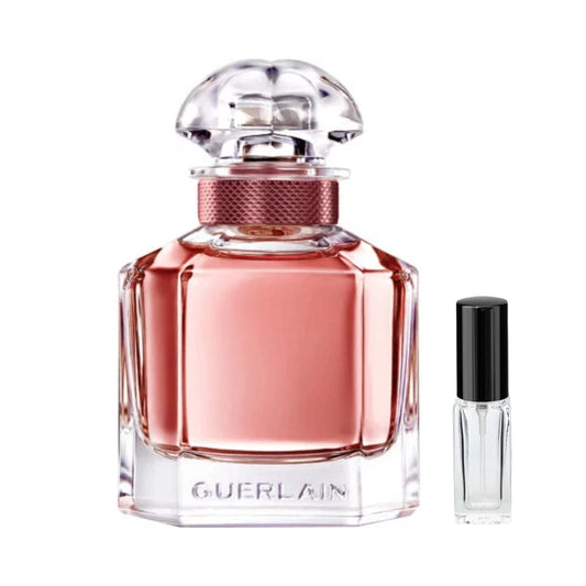 Szklany, fasetowany flakon damskich perfum Mon Guerlain Eau de Parfum Intense wypełniony miedziano-różowym płynem, z przezroczystym korkiem i teksturowaną, bordowo-miedzianą opaską wokół szyjki, a obok niego szklana fiolka odlewki – ODVARO.
