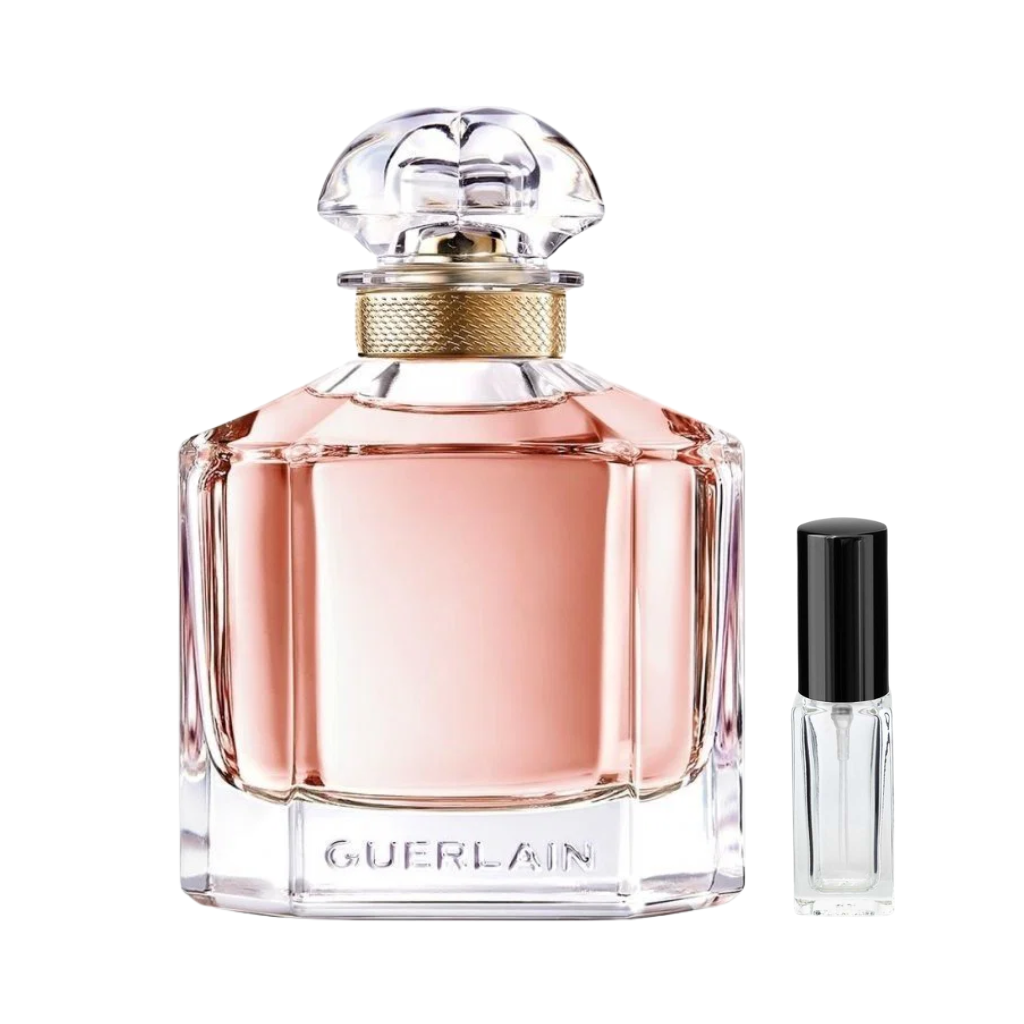 Przezroczysty, masywny flakon perfum Guerlain Mon Guerlain z różowym płynem, złotą plecioną obręczą na szyjce i przezroczystym korkiem, obok którego prezentowana jest szklana fiolka odlewki. – ODVARO