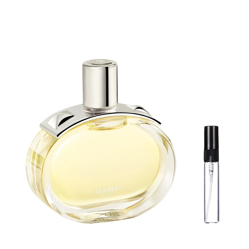 Owalny, szklany flakon perfum Hermès Barénia z jasnozłotym płynem, ozdobiony srebrną nakładką z detalem przypominającym ćwieki i gładkim, cylindrycznym korkiem, a obok widoczna szklana fiolka odlewki – ODVARO.