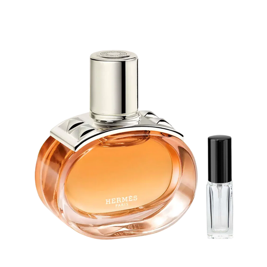 Owalny, przezroczysty flakon perfum Hermès Barénia Pleine Fleur z bursztynowym płynem i srebrnym korkiem z ćwiekami, obok którego znajduje się szklana fiolka odlewki – ODVARO.