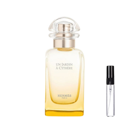 Smukły, prostokątny flakon perfum Hermès Un Jardin à Cythère o gradientowym, złocisto-żółtym szkle z czarnymi napisami i przezroczystym, półokrągłym korkiem, obok którego zaprezentowana jest szklana fiolka odlewki – ODVARO.