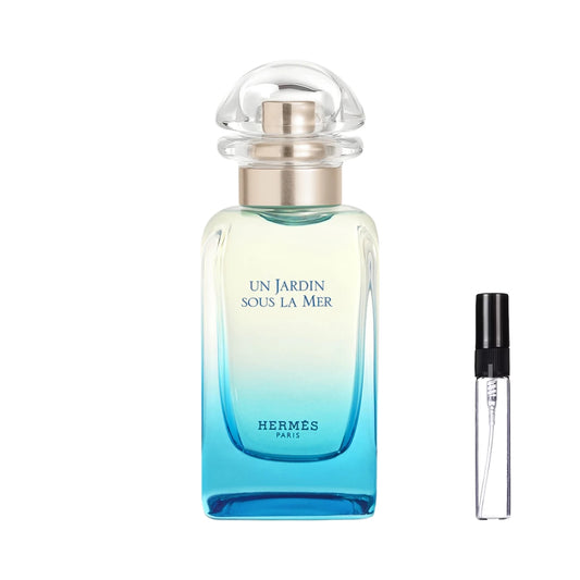 Smukły, prostokątny flakon perfum Hermès Un Jardin Sous la Mer o gradientowym szkle przechodzącym z delikatnej zieleni w głęboki, morski błękit z ciemnoniebieskimi napisami i przezroczystym, półokrągłym korkiem, obok którego zaprezentowana jest szklana fiolka odlewki – ODVARO.