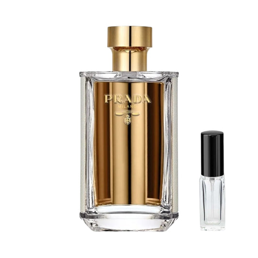 Szklany flakon damskich perfum Prada La Femme z eleganckim złotym wnętrzem, wyrazistym złotym logo Prada na froncie oraz klasycznym złotym korkiem, a obok niego szklana fiolka odlewki – ODVARO.