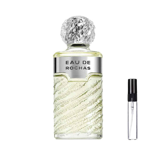 Szklany flakon perfum Eau de Rochas o fakturze przypominającej rzeźbioną skałę, z jasnozielonym płynem, szeroką srebrną etykietą i okrągłym, strukturalnym korkiem, obok którego zaprezentowana jest szklana fiolka odlewki – ODVARO.